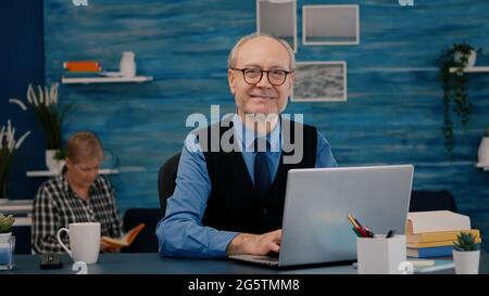 Pensionato manager seduto alla scrivania di fronte alla macchina fotografica sorridente dopo aver digitato al computer portatile lavorando da casa, mentre la moglie anziana leggere un libro in background. Uomo preparato per meeting online utilizzando gadget moden Foto Stock