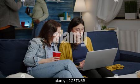Donne multietniche che si divertono insieme a guardare un film sul computer portatile mentre si riposano sul divano. In background donna e uomo bevono birra, ridendo a tarda notte durante la festa rilassante Foto Stock