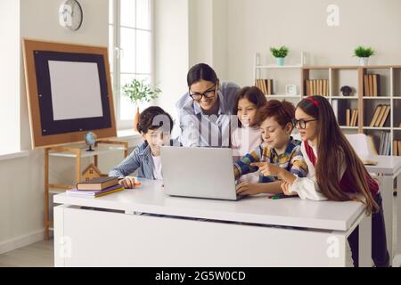 I bambini delle scuole elementari e le insegnanti di scuola elementare lavorano insieme su un computer portatile. Foto Stock