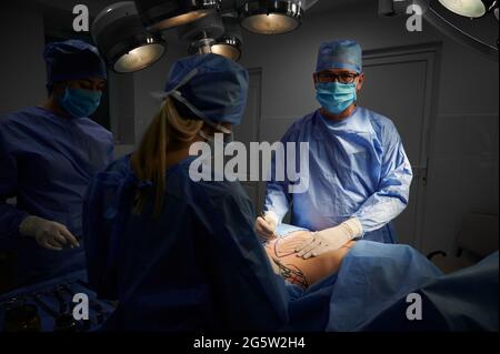 Team medico che si occupa di chirurgia plastica estetica in sala operatoria con luce debole. Chirurgo che esegue chirurgia estetica, guardando alla fotocamera. Concetto di medicina e chirurgia plastica Foto Stock