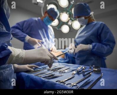 Primo piano di medico in piedi da strumenti medici in acciaio e pinzette di tenuta con tampone, mentre chirurgo e assistente che eseguono chirurgia plastica in sala operatoria. Concetto di strumenti medici. Foto Stock