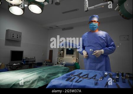 Ritratto di chirurgo in plastica che tiene in mano strumenti medici, in piedi in sala operatoria. Medico che indossa uniforme chirurgica, guanti sterili, maschera protettiva per il viso e cappuccio medico. Concetto di medicina Foto Stock