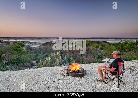 Camper seduto accanto al fuoco, guardando al lago artificiale Amistad al tramonto, campeggio Governors Landing, vicino a del Rio, Texas, USA Foto Stock