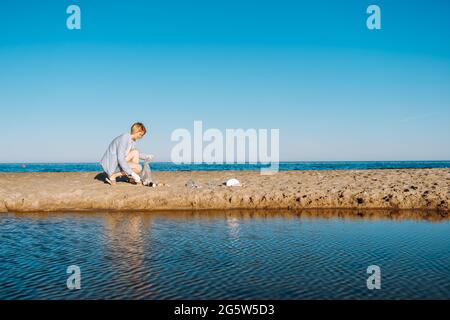 Una donna vestita con abiti casual raccoglie i rifiuti sulla spiaggia in una giornata estiva di sole. Spazio di copia. Zero sprechi. Foto Stock