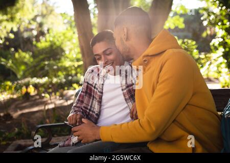Due amici maschi di razza felici seduti sulla panca del parco utilizzando lo smartphone Foto Stock