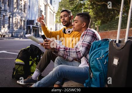 Due amici maschili di razza misti seduti in strada con i bagagli, utilizzando lo smartphone e guardando la mappa Foto Stock