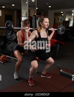 Allenatore personale femminile che aiuta la giovane donna che fa squats barbelll in palestra Foto Stock