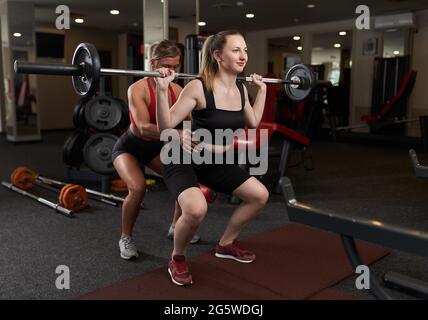 Allenatore personale femminile che aiuta la giovane donna che fa squats barbelll in palestra Foto Stock