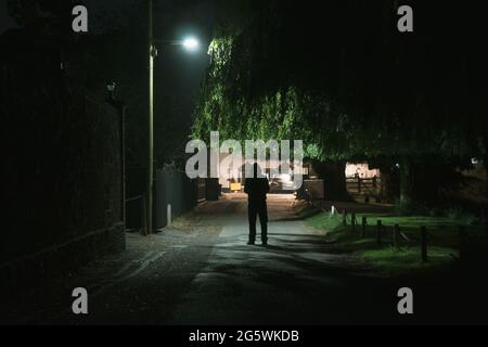 Una misteriosa figura incappucciata. In piedi sotto una luce di strada in una città. Di notte. Foto Stock