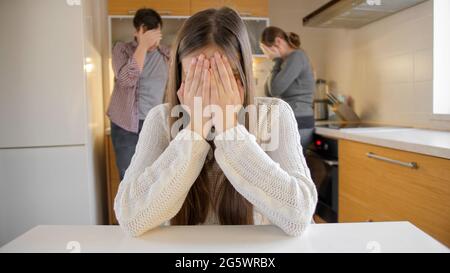 Ragazza sconvolto piangendo e chiudendo faccia con le mani perché su discutere e gridare i genitori in cucina. Violenza familiare, conflitti e problemi di relazione Foto Stock