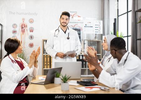 Studenti multirazziali in camici bianchi seduti in classe interattiva e aggrappati alla fine di una lezione interessante. Insegnante indiano che usa un computer portatile moderno per il processo educativo. Foto Stock