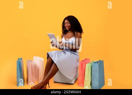 Lunghezza intera di donna nera graziosa con i sacchetti del regalo che usano il calcolatore del tablet per comprare le cose in linea sullo sfondo arancione dello studio Foto Stock