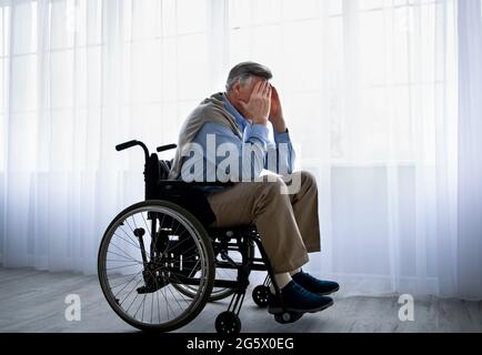 Uomo anziano portatore di handicap in sedia a rotelle coprendo il viso con le mani, piangendo vicino alle finestre, soffrendo di depressione all'interno Foto Stock