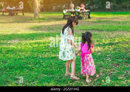 Vista posteriore delle bambine asiatiche che camminano fianco a fianco attraverso il giardino verde. Sorella maggiore e sorella minore che camminano insieme nel parco. Buona spesa per la famiglia Foto Stock