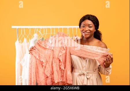 Stilista femminile professionista che tiene la blusa alla moda vicino al portabiti, scegliendo un abito elegante su sfondo arancione Foto Stock