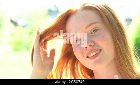 Ritratto una giovane donna sorridente con capelli rossi e fracelle sullo sfondo di un prato luminoso e soleggiato. Bellezza naturale con frettole Foto Stock