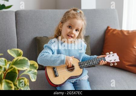 Ritratto di carina bambina che gioca ukulele e lo canta a casa. 3 anni bambino imparare chitarra. Concetto di educazione della prima infanzia, hobbistica musicale Foto Stock