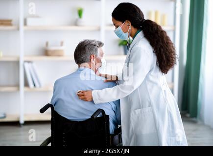 Giovane dottoressa in maschera facciale che ascolta la respirazione dell'anziano, usando stetoscopio a casa Foto Stock