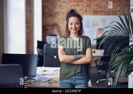 Ritratto di donna felice, designer di interni o architetto sorridente alla macchina fotografica mentre in piedi con le braccia incrociate in un moderno ufficio verde di giorno Foto Stock