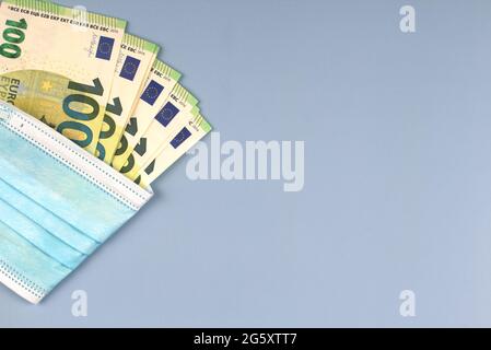 Un centinaio di banconote in euro avvolte in maschera chirurgica monouso su fondo grigio chiaro. Vista dall'alto Foto Stock