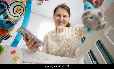 Giovane madre impegnata che scuote il giocattolo colorato per il suo bambino mentre usa lo smartphone. Concetto di parenting, i genitori occupati di lavoro e sviluppo del bambino Foto Stock