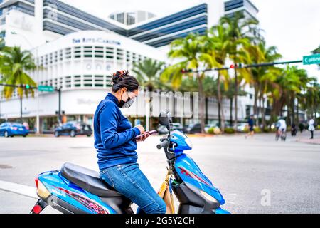 Miami Beach, USA - 17 gennaio 2021: Famoso Lincoln Road Collins Avenue intersezione e donna candida cavalcare su moto indossare maschera viso utilizzando mas Foto Stock