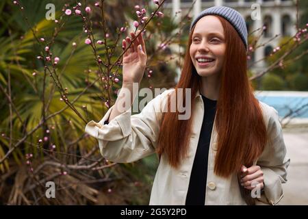 Sorprendente giovane donna dai capelli rossi in cappello che si pone in fiore albero a primavera. Bella giovane donna felice godendo l'odore nel giardino fiorente della molla, da sola, Foto Stock