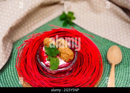 Parfait da dessert con marmellata di fragole in un vaso di vetro decorato con biscotti crackizzati, foglie di menta su un tovagliolo verde vimini. Una piccola porzione di cibo domestico. Foto Stock