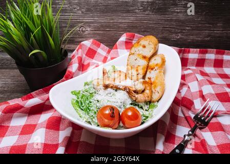 Insalata di gamberi in un recipiente con crostini croccanti, pomodori ciliegini, salsa bianca, formaggio grattugiato. Su un banco da cucina, una forchetta e un tovagliolo a scacchi Foto Stock