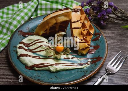 Waffle belgi fatti in casa e soffici con bacca di fisalis, una tazza di gelato e salsa al cioccolato in cima. Su un banco da cucina, tovagliolo a scacchi e asciutto Foto Stock