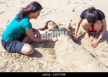 Belle donne indiane seppelliscono il loro grazioso amico nella sabbia sulla spiaggia, Puducherry (Pondicherry), Tamil Nadu, India Foto Stock