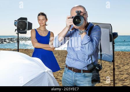 Ritratto di fotografo professionista che guarda attraverso l'obiettivo della fotocamera durante le riprese sulla spiaggia soleggiata Foto Stock