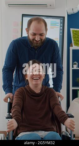 Giovane uomo allegro che spinge la sedia a rotelle con il suo collaboratore invalido paralizzato mentre sorride in start up ufficio finanziario di affari. I colleghi si divertono a ridere sul posto di lavoro durante il lavoro Foto Stock