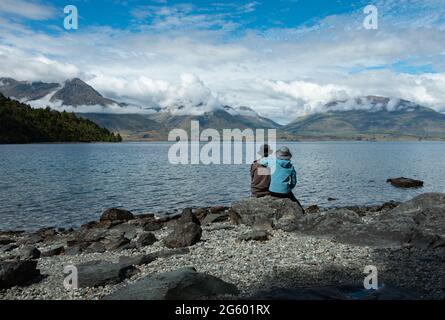 Una coppia seduta sulle rocce, godendo la vista sulle montagne attraverso il lago Wakatipu, Queenstown. Foto Stock