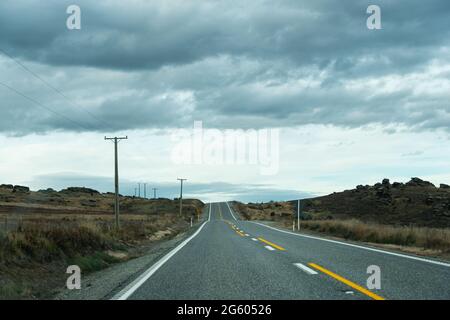 Una strada di campagna vuota con linee elettriche lungo la strada e nuvole scure in testa, la regione di Otago, Isola del Sud, Nuova Zelanda Foto Stock