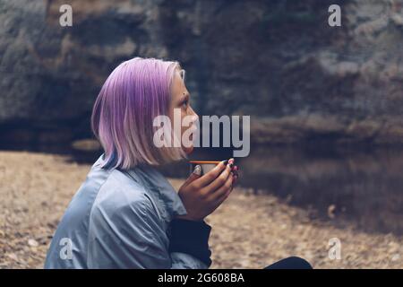 ritratto di una ragazza adolescente che riposa con una tazza sulla riva del fiume guardando le rocce costiere Foto Stock