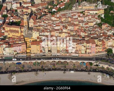 VISTA AEREA. Old Town con case colorate a più piani che scende lungo una ripida collina verso la strada fronte spiaggia. Menton, Costa Azzurra, Francia. Foto Stock