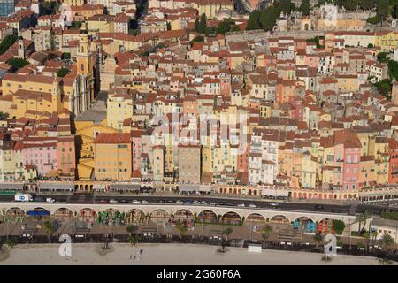 VISTA AEREA. Old Town con case colorate a più piani che scende lungo una ripida collina verso la strada fronte spiaggia. Menton, Costa Azzurra, Francia. Foto Stock