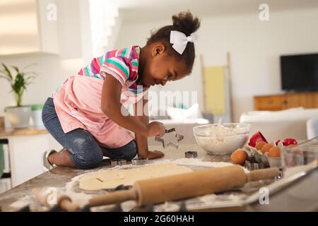 Felice figlia afroamericana inginocchiata in cucina contro taglio forme in pasta Foto Stock