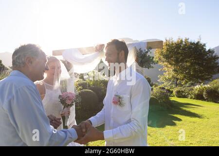 Felice sposa caucasica e sposo sposarsi scuotendo le mani con officiant matrimonio Foto Stock