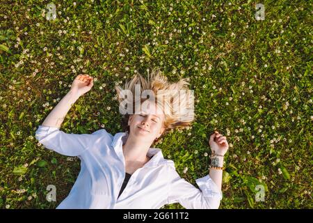 Vista dall'alto di una giovane donna sdraiata su un prato con trifoglio fiorito a occhi chiusi. Insetti, allergie, acari primaverili Foto Stock