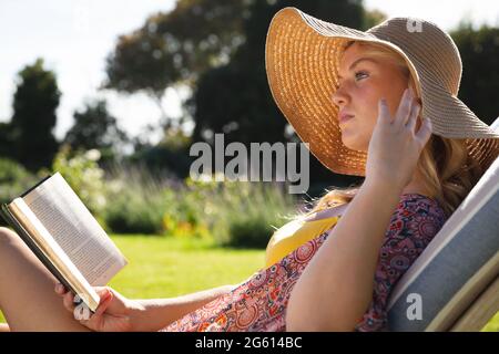 Donna caucasica che indossa cappello rilassante sul lettino in giardino soleggiato lettura libro Foto Stock