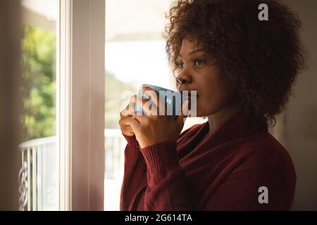 Donna afro-americana pensierosa in piedi alla finestra soleggiata bere caffè e sorridente Foto Stock