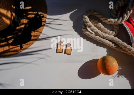 Borsa da spiaggia, occhiali da sole, bikini, cappello di paglia, orecchini, frutta arancione in foglie di palma ombra su sfondo bianco. Appartamento estivo posato. Vacanza concep Foto Stock