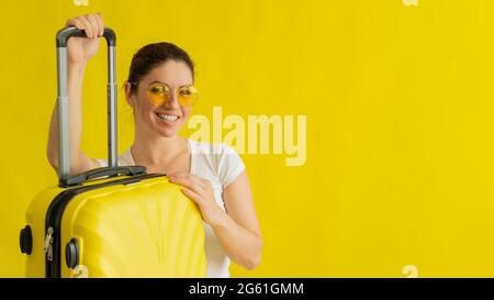 Donna sorridente in occhiali da sole con valigia con manico retrattile su sfondo giallo Foto Stock