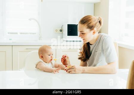 Giovane bella madre nutre il bambino con un cucchiaio di purea di verdure in cucina. Foto Stock