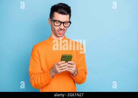 Ritratto di giovane ragazzo strano tenere la lingua del telefono fuori dispiaciuto occhiali isolati su sfondo di colore blu Foto Stock
