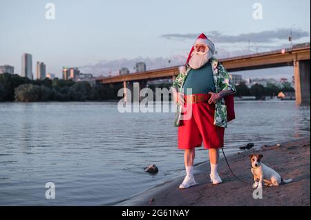 Babbo Natale indossa pantaloncini che camminano lungo la spiaggia con il cane Jack Russell Terrier. Foto Stock