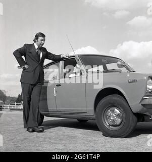 1975, su una zona asfaltata fuori strada, un uomo in tuta che parla con una donna attraente in occhiali da sole seduto nella sua nuova Datsun Cherry 100A auto, Inghilterra, Regno Unito. Si noti l'ampio lapel della giacca da uomo e i pantaloni svasati dei pantaloni e le scarpe con plateau, stili di moda distintivi degli anni '70. Nissan ha iniziato ad esportare nel Regno Unito automobili con logo Datsun nel 1968 e il marchio giapponese ha avuto un grande successo, con auto affidabili e a buon prezzo, in un momento in cui l'industria automobilistica britannica era in crisi, con scioperi da parte della forza lavoro e una reputazione per la realizzazione di auto di scarsa qualità e affidabilità. Foto Stock