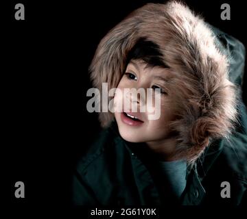 ragazzo con un aspetto tenero che guarda nel profilo sorridente con un caldo parka in pelliccia cappuccio, orizzontale sfondo nero e spazio di copia Foto Stock